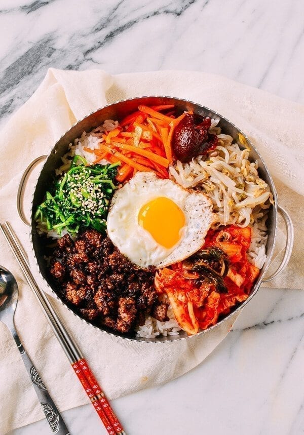 Top-down viw of bibimbap in a metal pot, showing a fried egg, beef, and various vegetables. A spoon and pair of metal chopsticks are to the left.