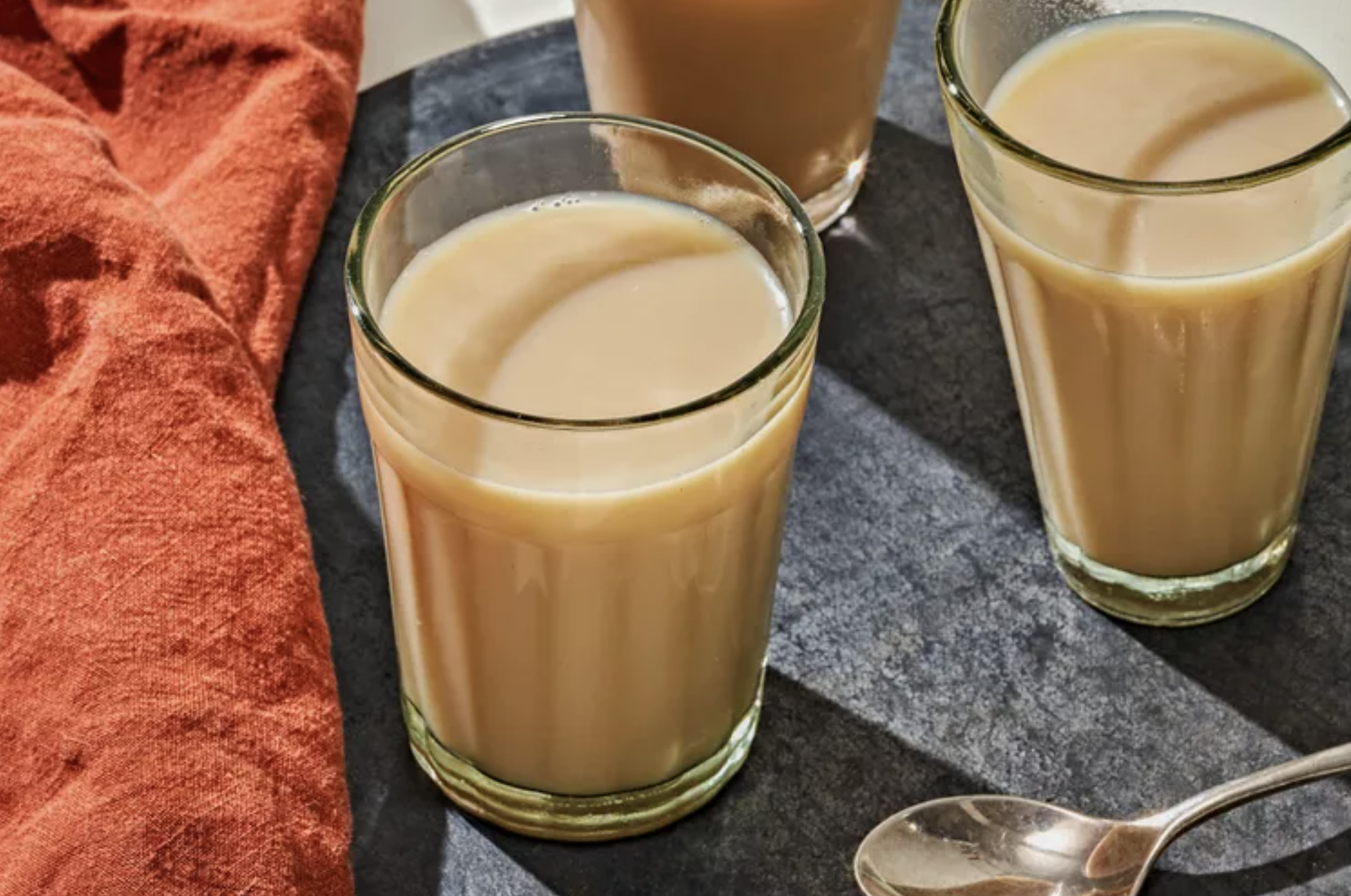 Two glasses of chai on a gray surface, with some orange fabric to the left and a spoon in front.